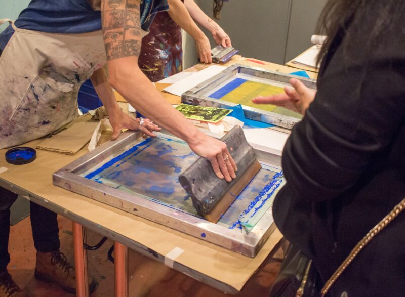 Two individuals silkscreen printing a camouflage image in blue while another person observes.