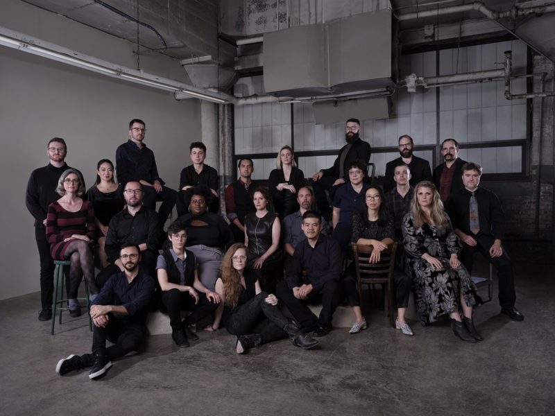 A large group of people sitting together in a large, warehouse-like room.