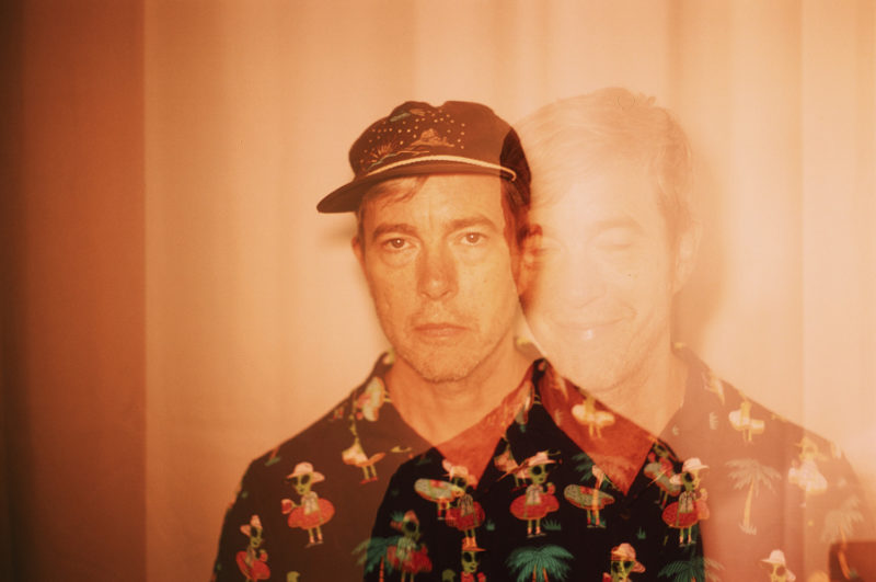 Double exposed photo of Bill Callahan in a button down shirt with vacationing aliens and palm trees on it. In one of the exposures he is wearing a hat and looking stoically at the camera. In the other exposure, he is not wearing a hat and is smiling.
