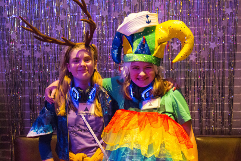 Two people with medium length hair stand in front of a silver streamer backdrop while they have their arms around each other, posing for a photo. One is wearing a colorful court jester hat and the other is wearing antlers. They each have headphone sets with blue lights around their necks.
