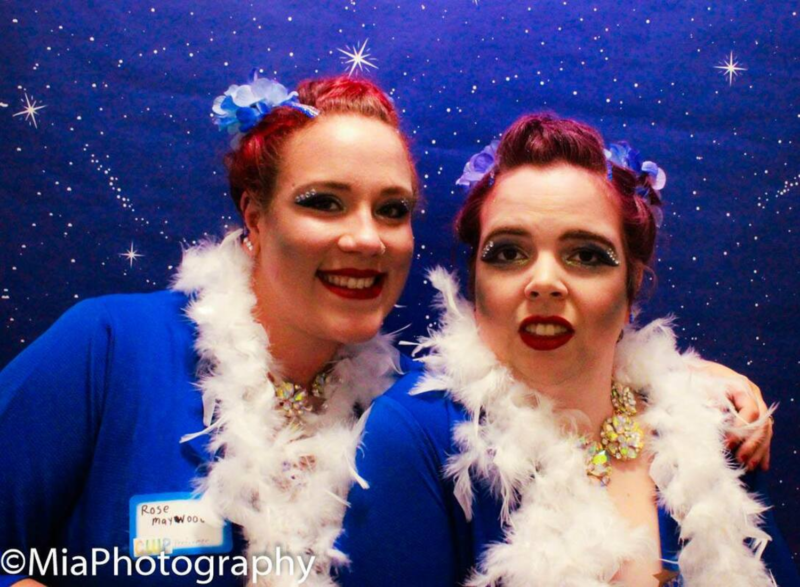 Two people posing in front of a blue night sky background. They are smiling and wearing blue tops with white feather boas wrapped around their necks. They each have flowers in their red hair and have rhinestones on their eye makeup.