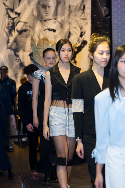 A group of teens are walking in a line towards the right side of the camera in the entrance space of The Andy Warhol Museum.