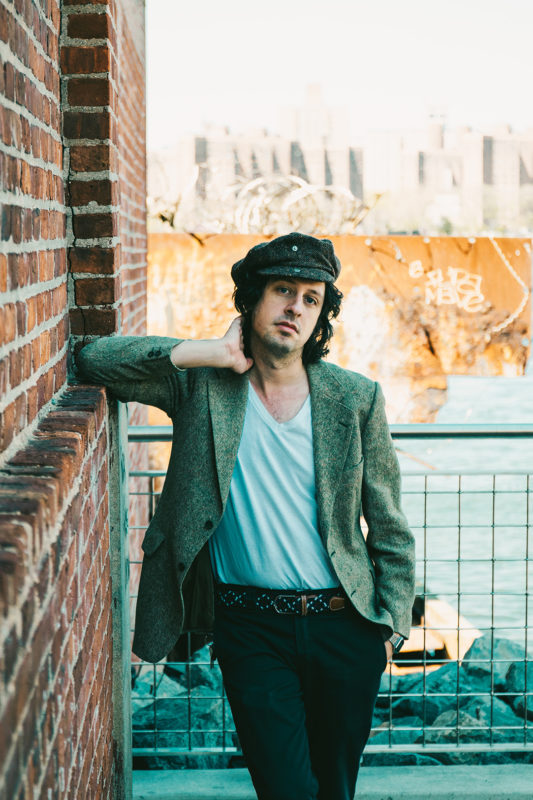 Adam Green is standing outside leaning against a red brick building with his right arm. He is wearing a black hat, light colored t-shirt, gray sport coat, and dark colored pants.