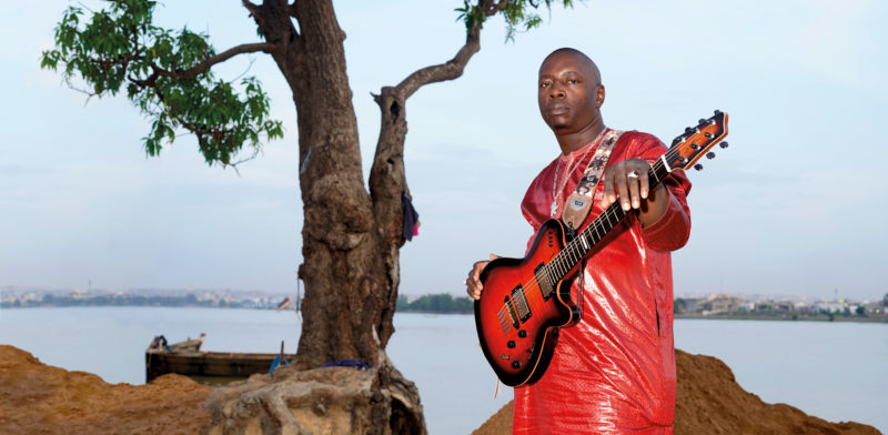 Vieux Farka Touré stands near the edge of a body of water near a tree. He is wearing a red outfit and holding a red and black guitar.