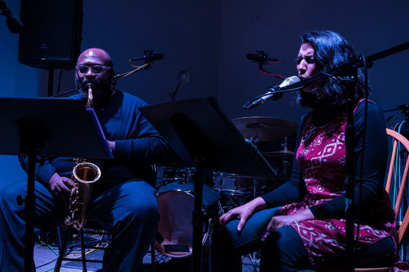 Amirtha Kidambi and Darius Jones are sitting on chairs in a room with a drumset behind them. In front of them are music stands and microphones. Darius is playing a saxophone and Amirtha is singing into the microphone.