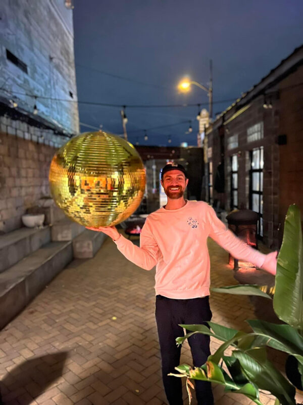 DJ Jarrett Tebbets stands outside in a pink sweatshirt holding a giant gold ball in his right hand.
