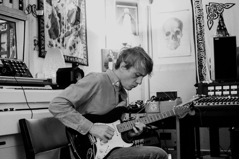 Black and white photograph of Steve Gunn sitting down playing a guitar in a room with recording equipment.