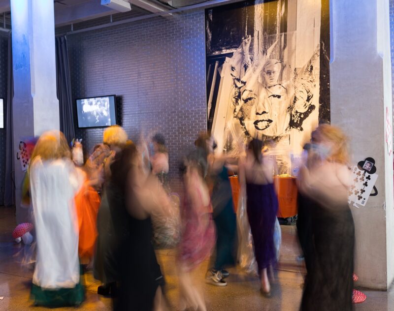 A group of people dancing causing them to look blurry. On the wall behind the group of people, a picture of Andy Warhol holding up a large acetate of Marilyn Monroe’s face.