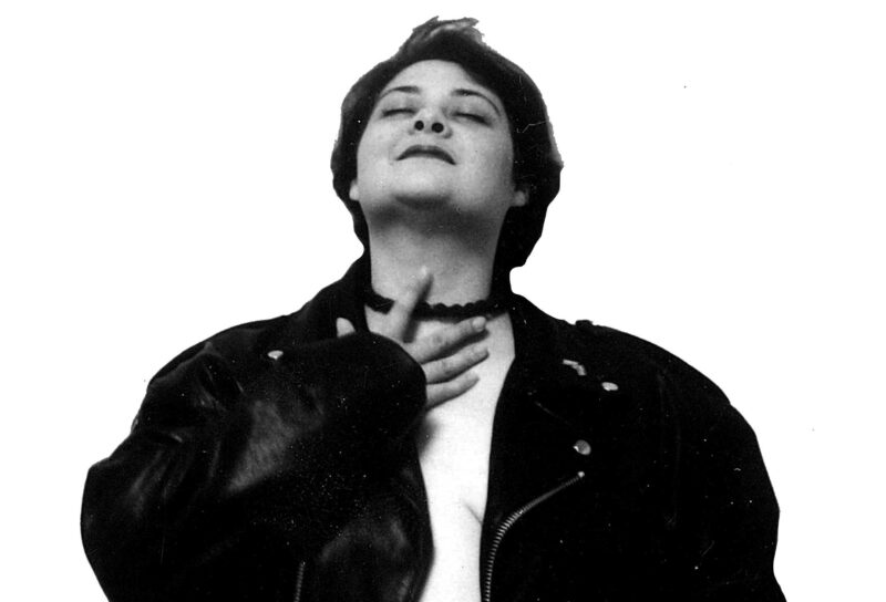 Image of woman in black and white. They are wearing a leathe rjacket and gazing upwards while her hand touches her chest.