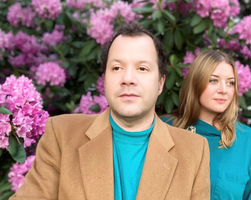 A man and woman both wearing teal shirts standing in a group of pink flowers. The man is also wearing a tan sport jacket.