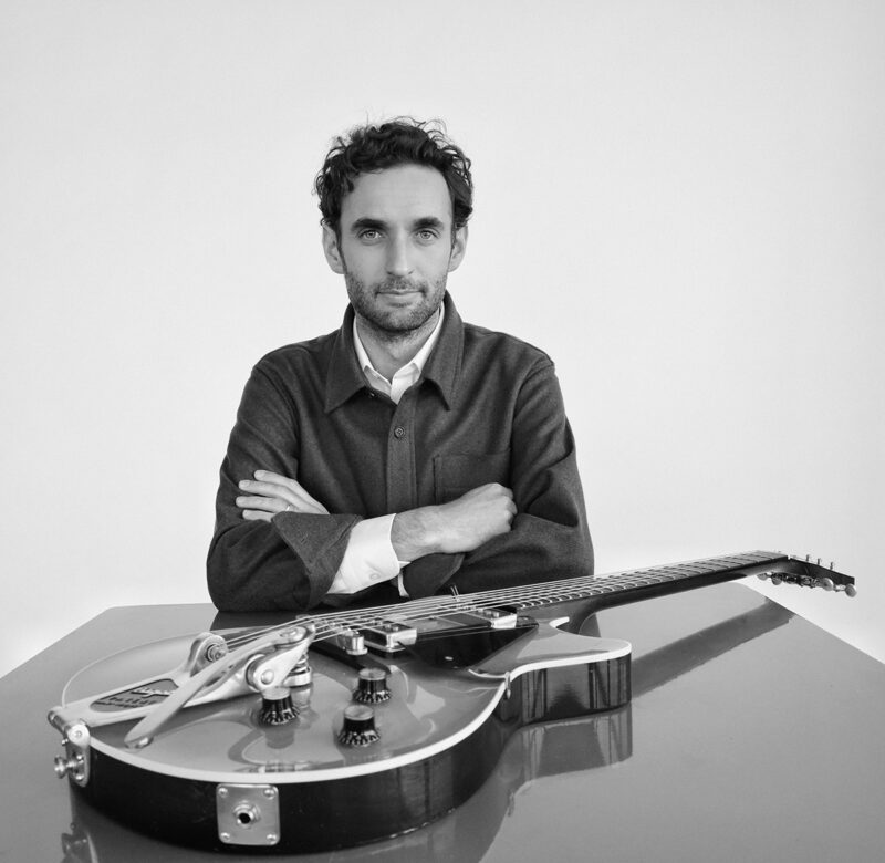 Black and white photo of Julian Lage sitting at a table in front of a white background with a guitar laying on the table in front of him.