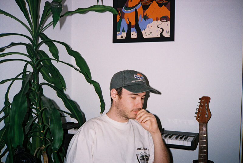 Elijah Wolf sits in a room in a white t-shirt and gray hat. There is a keyboard, guitar, artwork, and green plant in the background.
