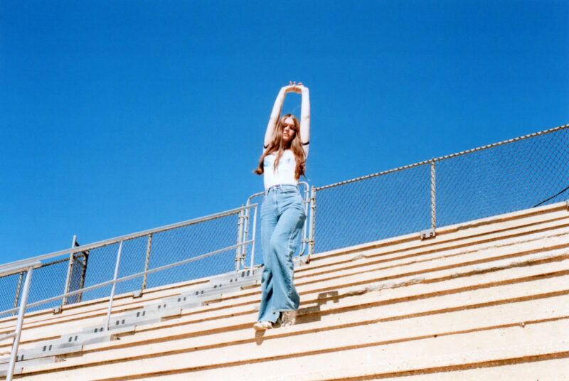 Ethel Cain stands on bleachers outside in the sun with her hands stretched up into the air.