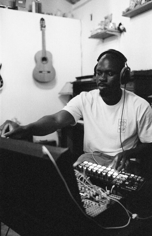 Black and white photograph of Colloboh wearing headphones while sitting at a desk in front of a monitor playing electronic instruments.