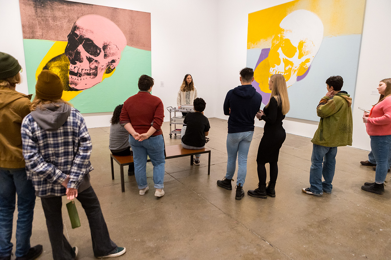 A group of people are in a gallery in The Andy Warhol Museum with two large screenprints of Warhol's "Skull" series hanging on the walls. The people are facing one person who appears to be giving a tour.