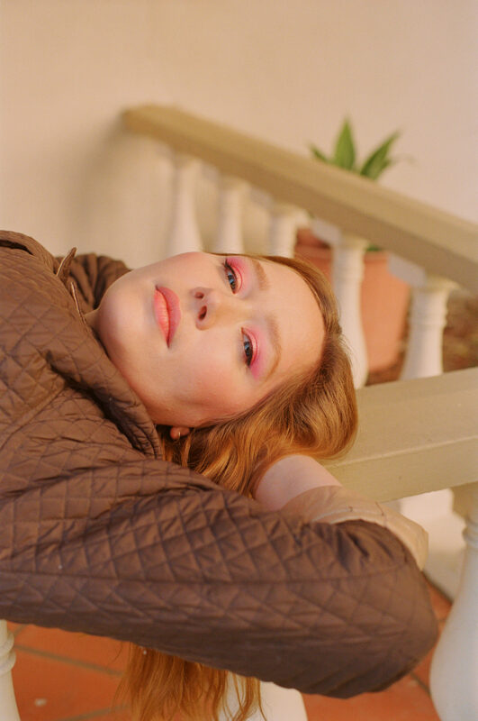 Julia Jacklin taken from the chest, up. She is laying on a railing with her hands behind her head smiling towards the camera.