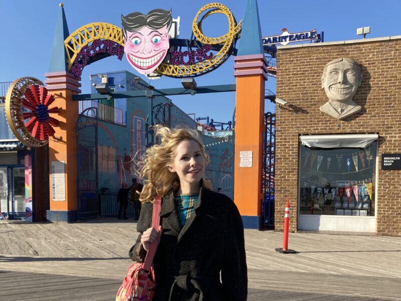 Nellie McKay is standing outside on a boardwalk in front of a carnival-like place that has a sign that looks like a clown face with Scream Zone around it. She is wearing a black coat and holding a large, pink purse.