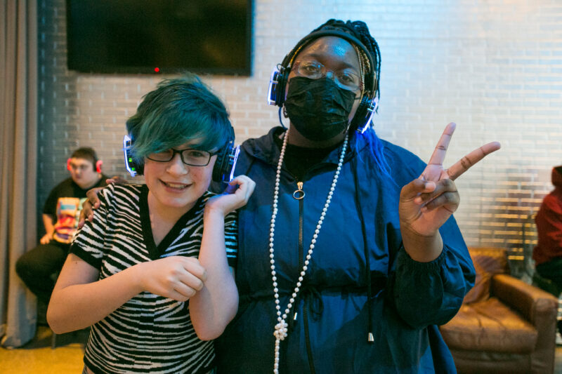 Two people pose for the camera wearing headphones with neon lights. One is wearing a face mask and giving the “peace” sign with their left hand and their right arm is around the other person. The other person is wearing a striped shirt and glasses and smiling for the camera.