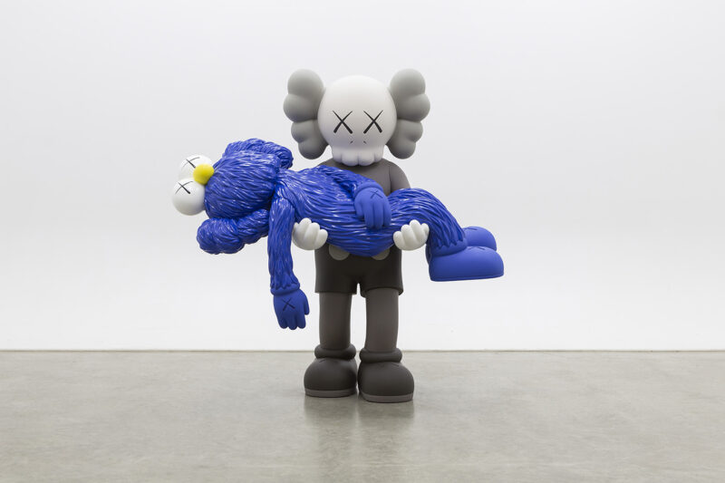 A black and white figure sculpture with x's on their eyes carries a blue sculpture that appears to have fur with x's on their eyes and a yellow nose.