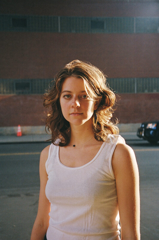 Allegra Krieger is wearing a white tank top and has shoulder length brown hair. She is standing outside across the street from a red brick building and looking towards the camera.