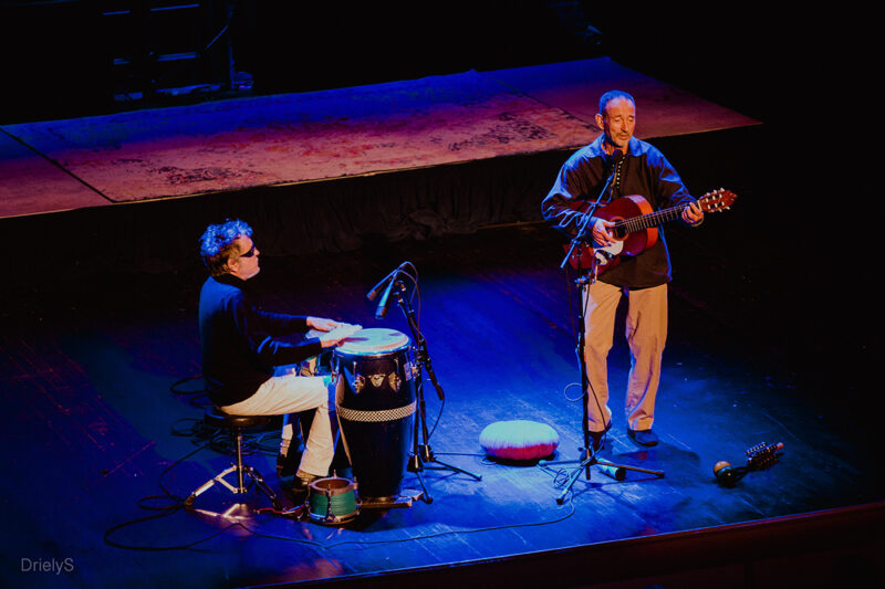 Johnathan Richman is standing on a stage singing into a microphone with a guitar in his hand. There is a person to his right on the stage playing bongo drums.