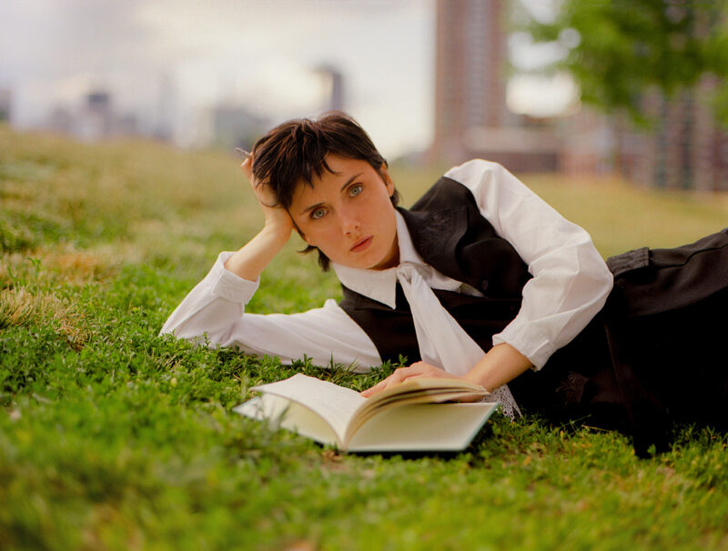 Katy Kirby lays outside on grass with a book open in front of her.