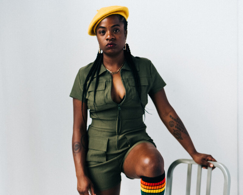 Sunny War stands in front of a white background with her left foot up on a metal chair. She is wearing a green jumpsuit and yellow berret.