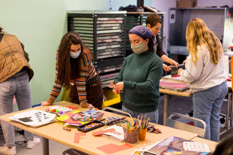 Two people are standing behind a table with arts supplies all over it in The Warhol's Factory studio. One person is leaning on the table while the other cuts pieces of paper.