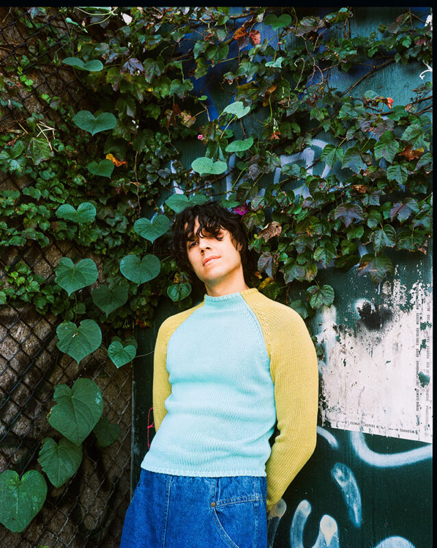 A person with dark, shoulder length hair, and wearing a blue sweater with yellow sleeves and jeans, is standing outside in the corner of a wall and fence that is covered in vines with green leaves.