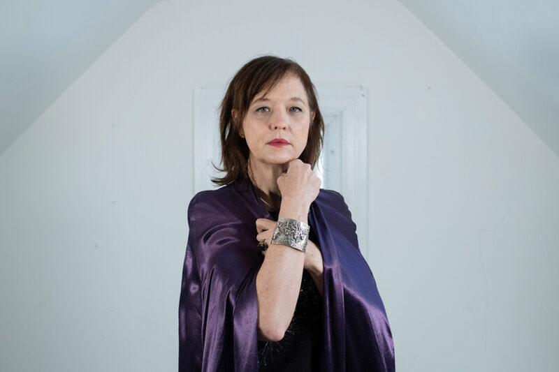 Mary Timony stands in a room with white walls. She is wearing a purple cape and is looking towards the camera with her right arm up across her chest.