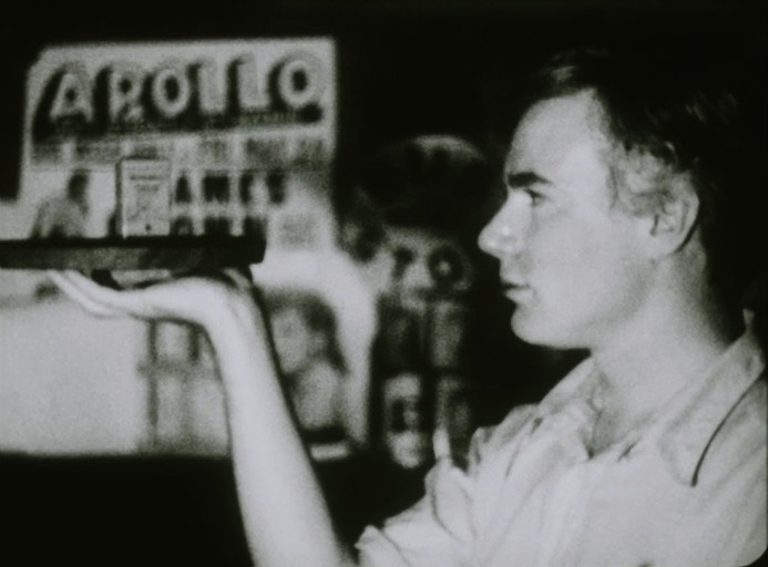 A man, Andy Warhol, holds a tray with one hand, his face is in profile with only his left side showing. In the tray is a small rectangular object. In the background the words Apollo can be made out.