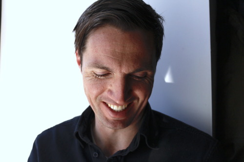 A close up of a person smiling and looking down in front of a white background.