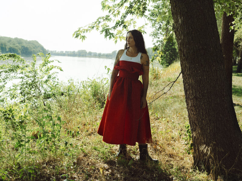 Person in a red and white dress, standing beside a tree along a river bank.