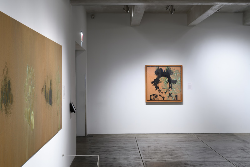 Installation view of the gallery. The painting on the left is half visible, a large rectangular brownish-gold abstract painting. The square painting on the adjacent wall is a silkscreened black portrait over a similar brownish-gold abstract painting.