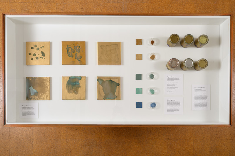 Overhead view of the gallery vitrine. The left side shows 6 small stretched canvases with brownish-gold abstract paintings. The right side shows 5 different colored samples, and 6 cylindrical containers.