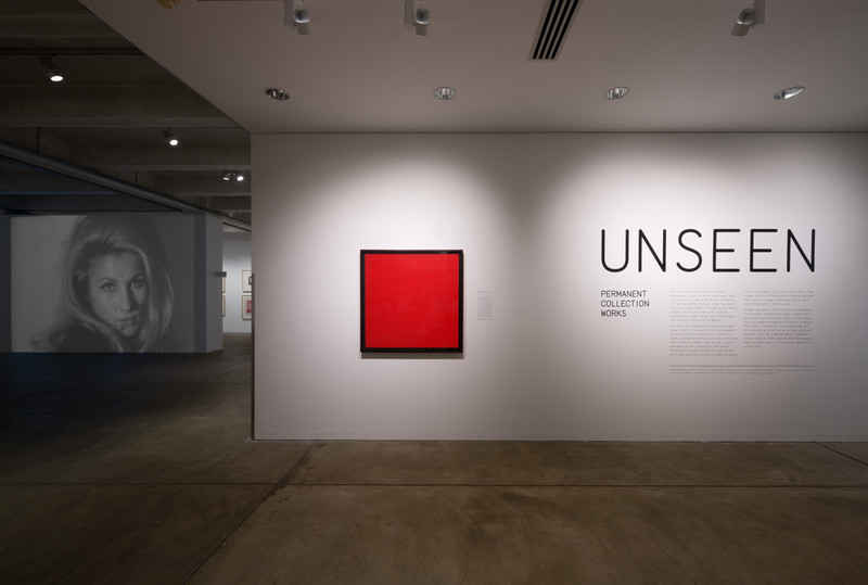 Installation view. Far left is a projection of a black and white film of a woman looking directly into the camera. The Center is a square painting that is completely red. And the far right is the exhibition wall text reading Unseen in large black letters.