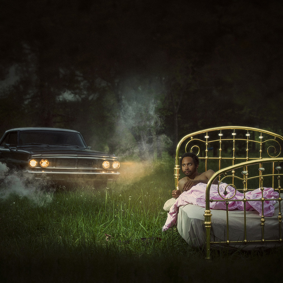 A man in a brass bed in a field at night with a car in the background.