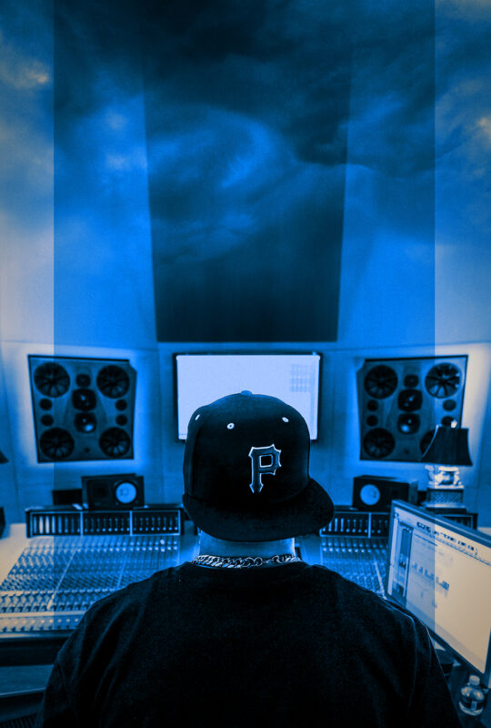 A photo taken behind a person sitting in front of a large audio mixing board. They are wearing a backwards Pittsburgh Pirates hat and a gold necklace.
