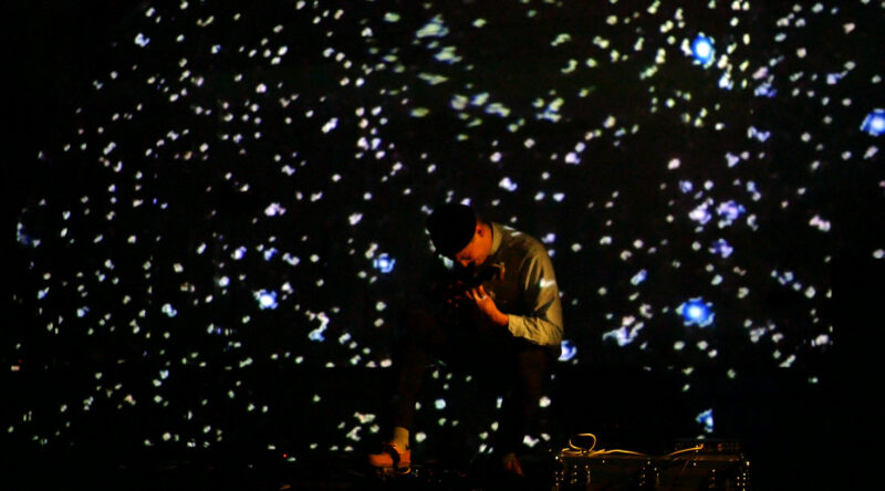 Patrick Higgins stands on a dark stage with small beams of light projected on it playing guitar.