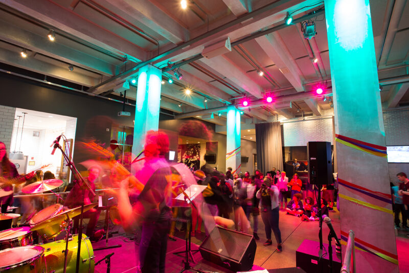 Blurry musicians in a band performing on a stage with the audience watching in the background lit with pink lighting.