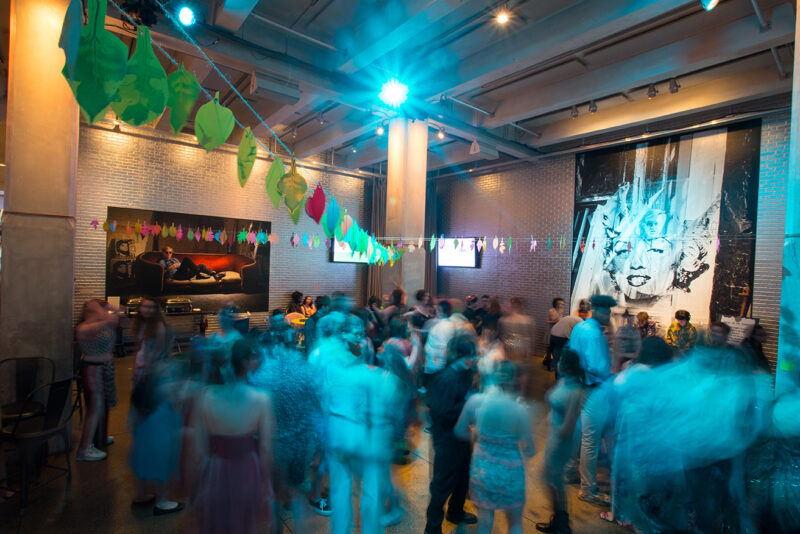 People fill the large open room of The Warhol’s entrance space. Colorful blue light covers their blurry figures, dancing and chatting excitedly under rainbow decorations.