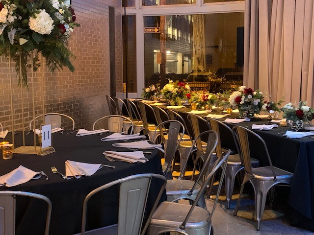 Two tables set for for a dinner one round smaller table and the other a long rectangular table both with black table cloths and flowers.