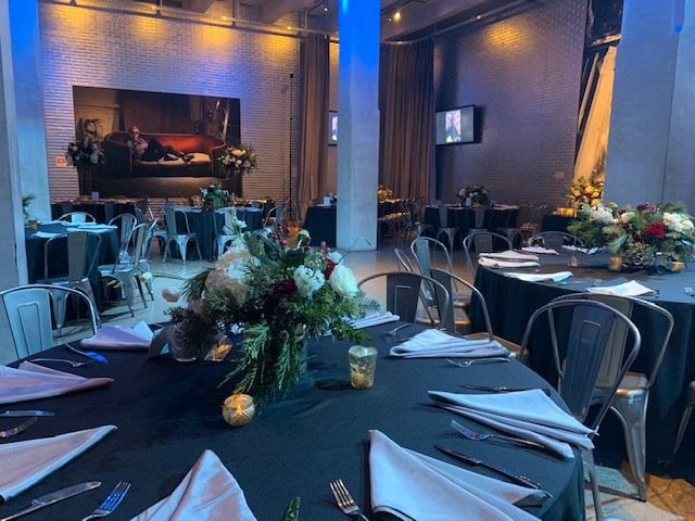 Several dinner tables are set with black table cloths, silver napkins, and flower centerpieces.