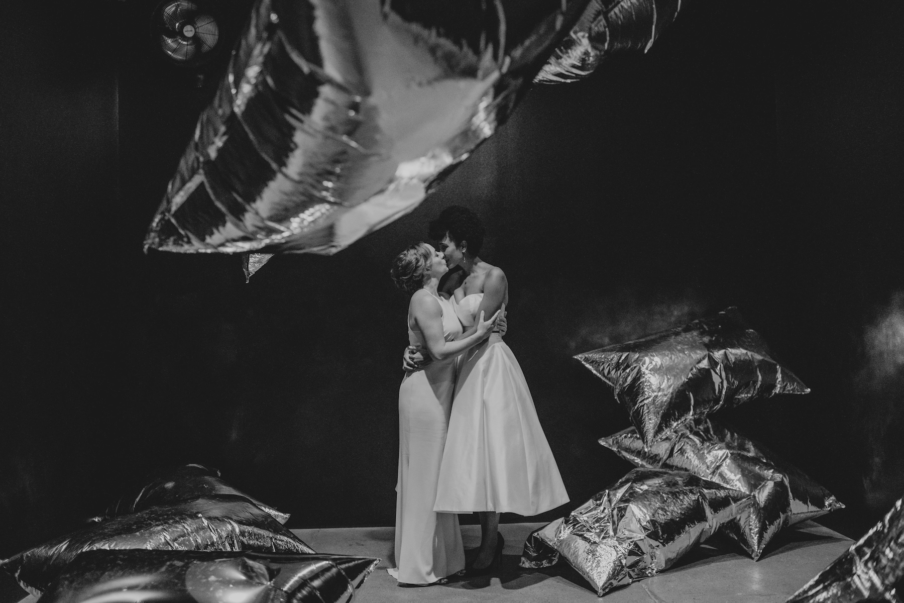 Black and white photography of two people embracing for a kiss, both wearing light dresses. They stand in the middle of a dark room with Andy Warhol's Silver Clouds (large mylar silver balloons) floating around them.