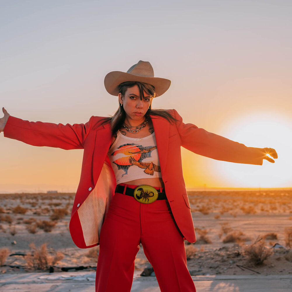 Alynda Segarra stands in a dessert as the sun is on the horizon looking towards the camera with her arms spread out. She is wearing a red suit with a tank top and tan cowboy hat.