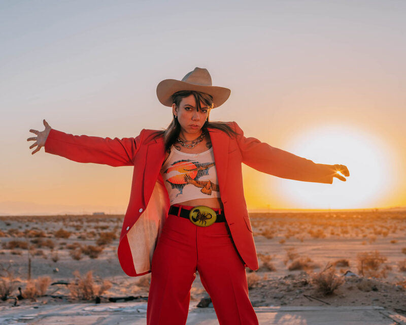 Alynda Segarra stands in a dessert as the sun is on the horizon looking towards the camera with her arms spread out. She is wearing a red suit with a tank top and tan cowboy hat.