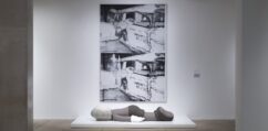 Painting showing two identical images of a car crash and body hanging out of a car window. In front of the painting a sculpture shows a figure laying face down.