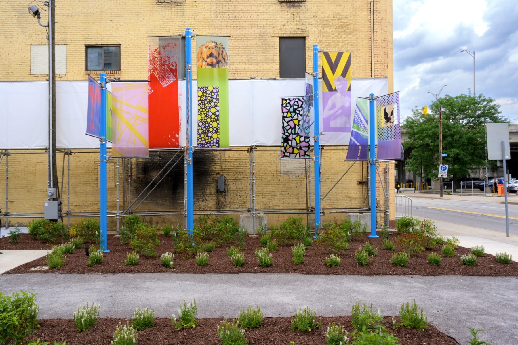 Installation image of art on 4 blue poles each with two colorful transparent banners that have various images on them.