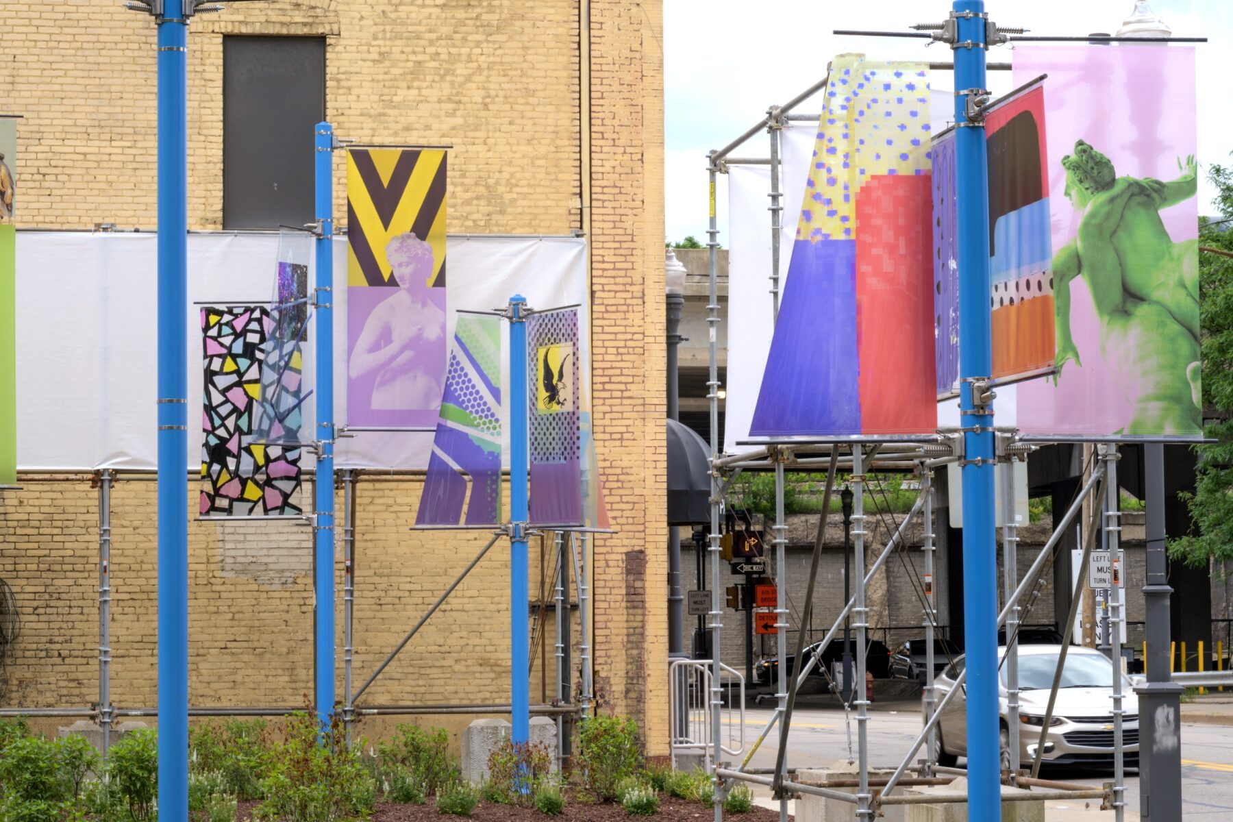 Installation image of art on 4 blue poles each with two colorful transparent banners that have various images on them.