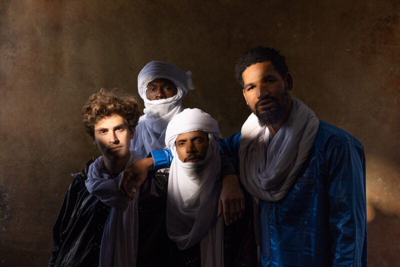 Four individuals wearing traditional Tuareg attire pose against a textured brown background. Mdou Moctar
